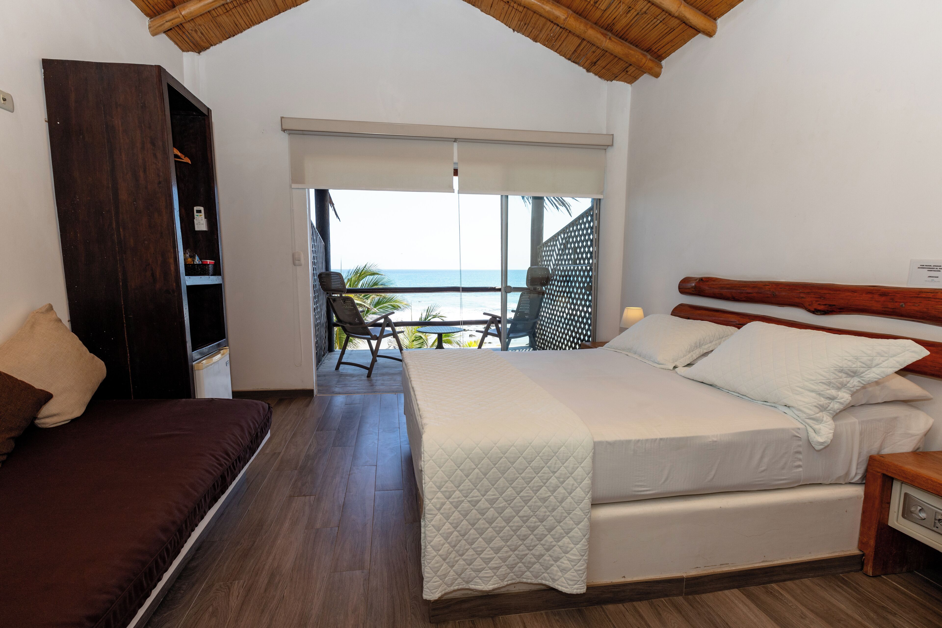 A bedroom with a bed, a wardrobe, a balcony with a view of the ocean, and a seating area with a table and chairs.