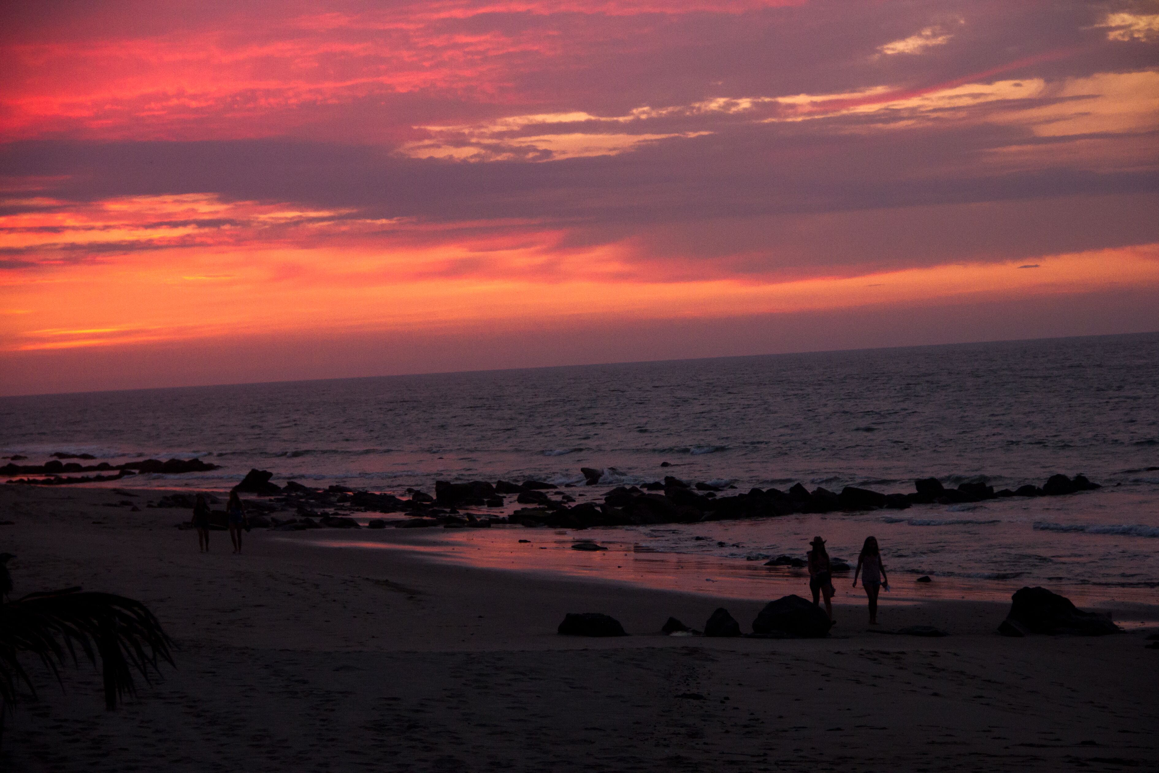Uma praia com pessoas caminhando pela orla, pedras na água e um céu ao pôr do sol.