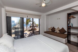 Un dormitorio con cama, un banco con cojines, ventilador de techo y vista a la playa y palmeras.