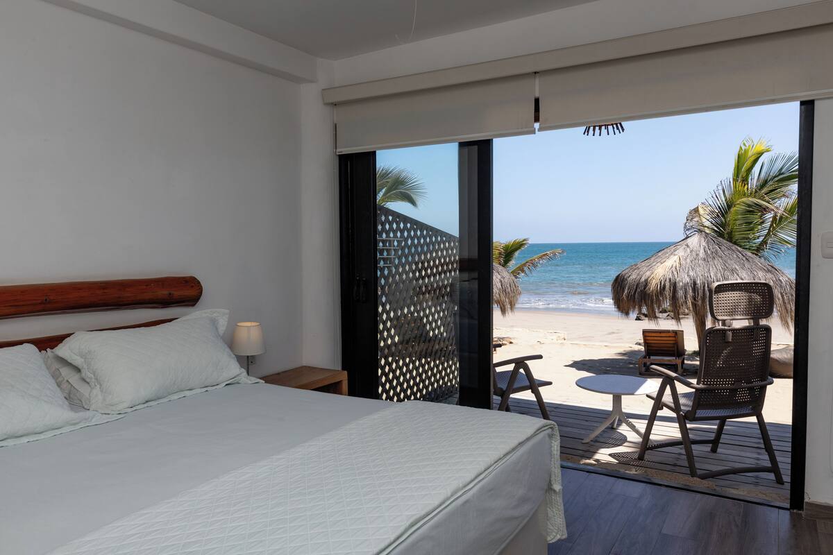 Ein Schlafzimmer mit Bett, Nachttisch und Blick auf einen Strand mit einer Reetdach-Sonnenschirm und Stühlen.
