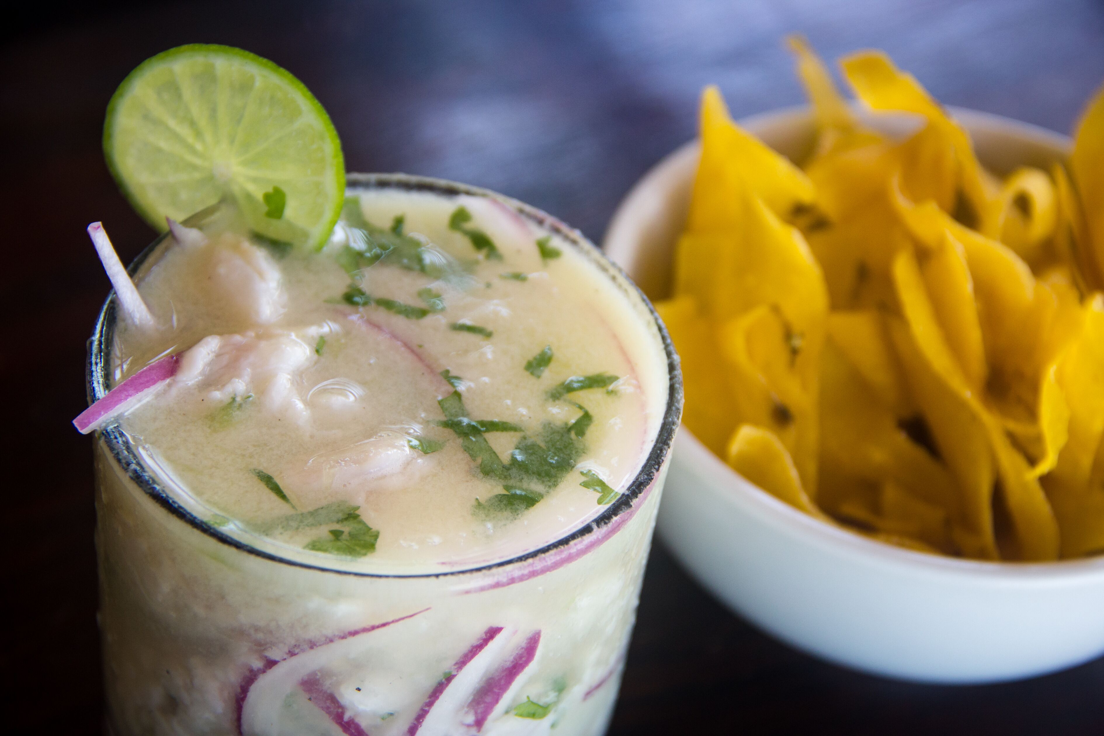 A glass of a creamy beverage with a lime slice garnish, accompanied by a bowl of yellow fruit slices.
