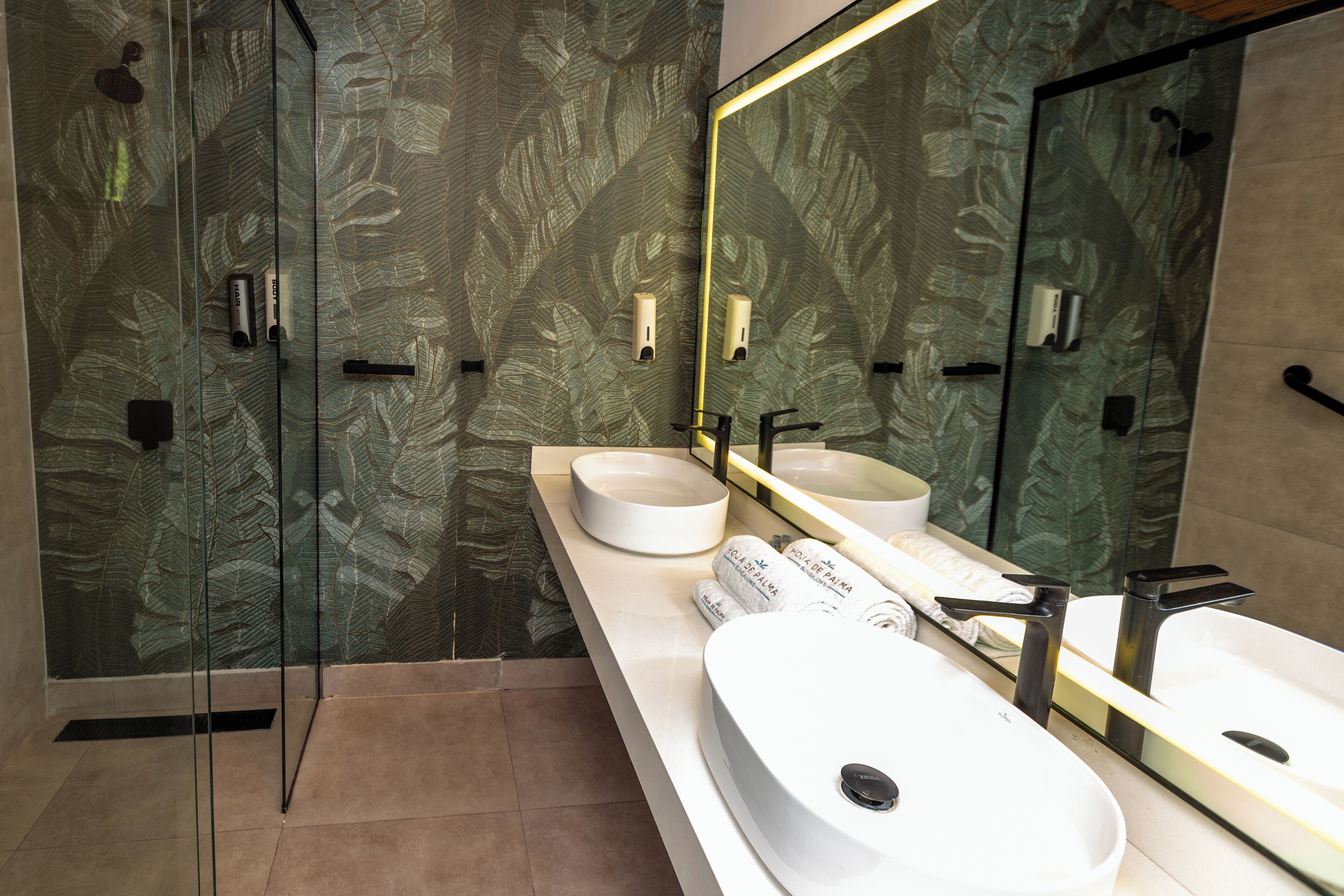 Une salle de bain moderne avec une douche à l’italienne en verre, un lavabo blanc avec un robinet noir et un grand miroir avec un cadre doré.