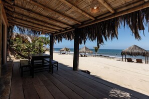 Una terraza de madera con techo de paja que da a una playa con sillones de descanso y palmeras.