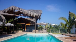 Un espace piscine agrémenté d’une structure à toit de chaume, de chaises longues et de parasols.