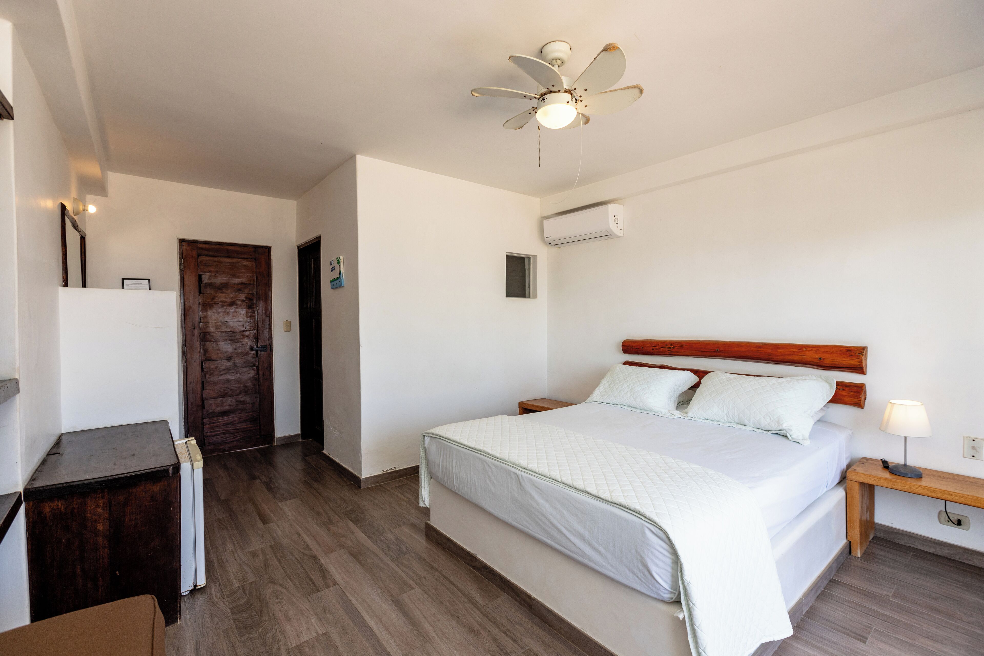 A neatly arranged bedroom with a wooden headboard, a single bed, a bedside table with a lamp, a ceiling fan, and an air conditioning unit.