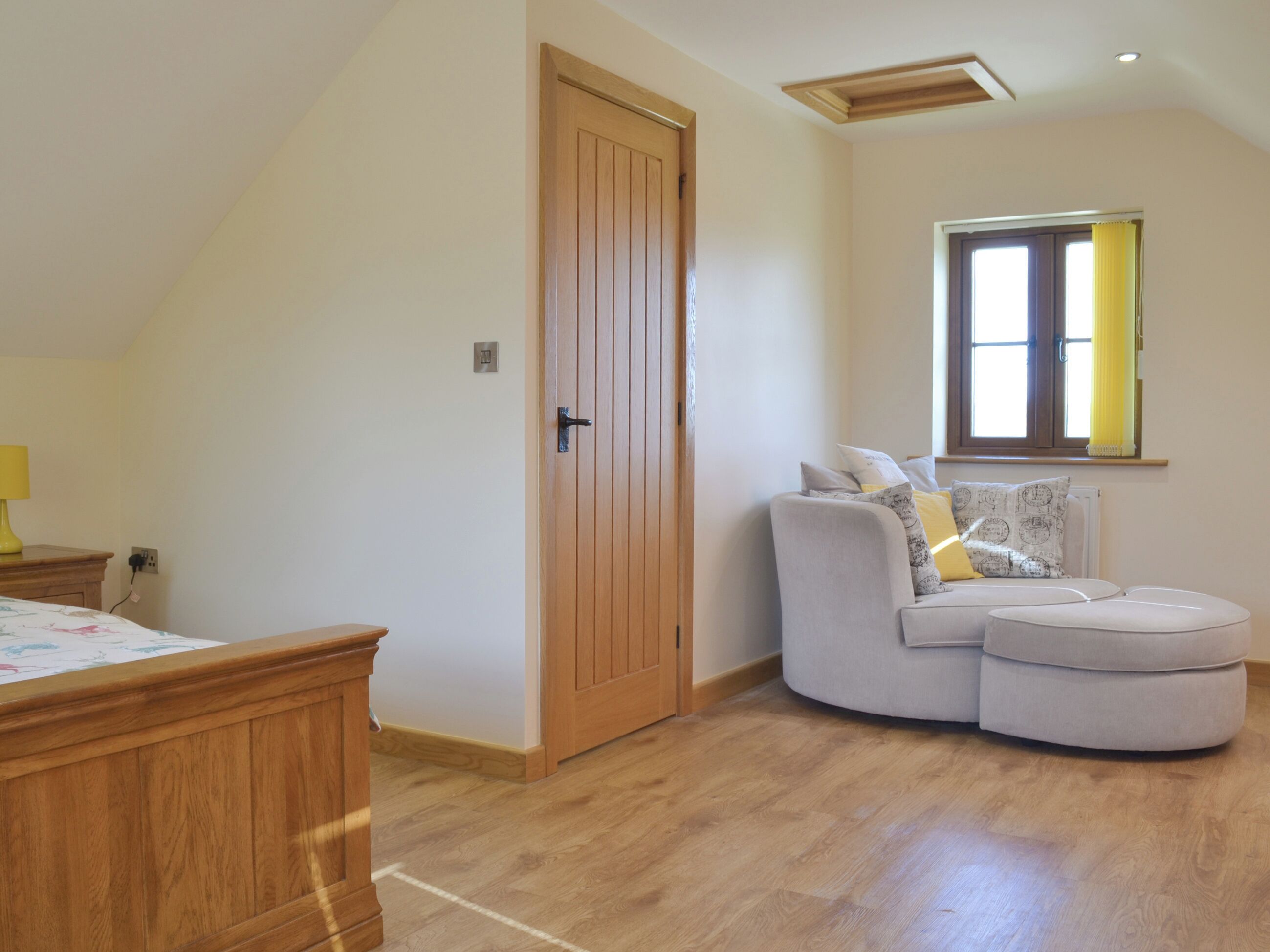A room with a wooden door, a curved grey sofa with cushions, a wooden bedside table, and a window with yellow curtains.