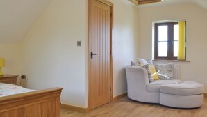 A room with a wooden door, a curved grey sofa with cushions, a wooden bedside table, and a window with yellow curtains. - 2 bedroom accommodation in Chard (Chard)