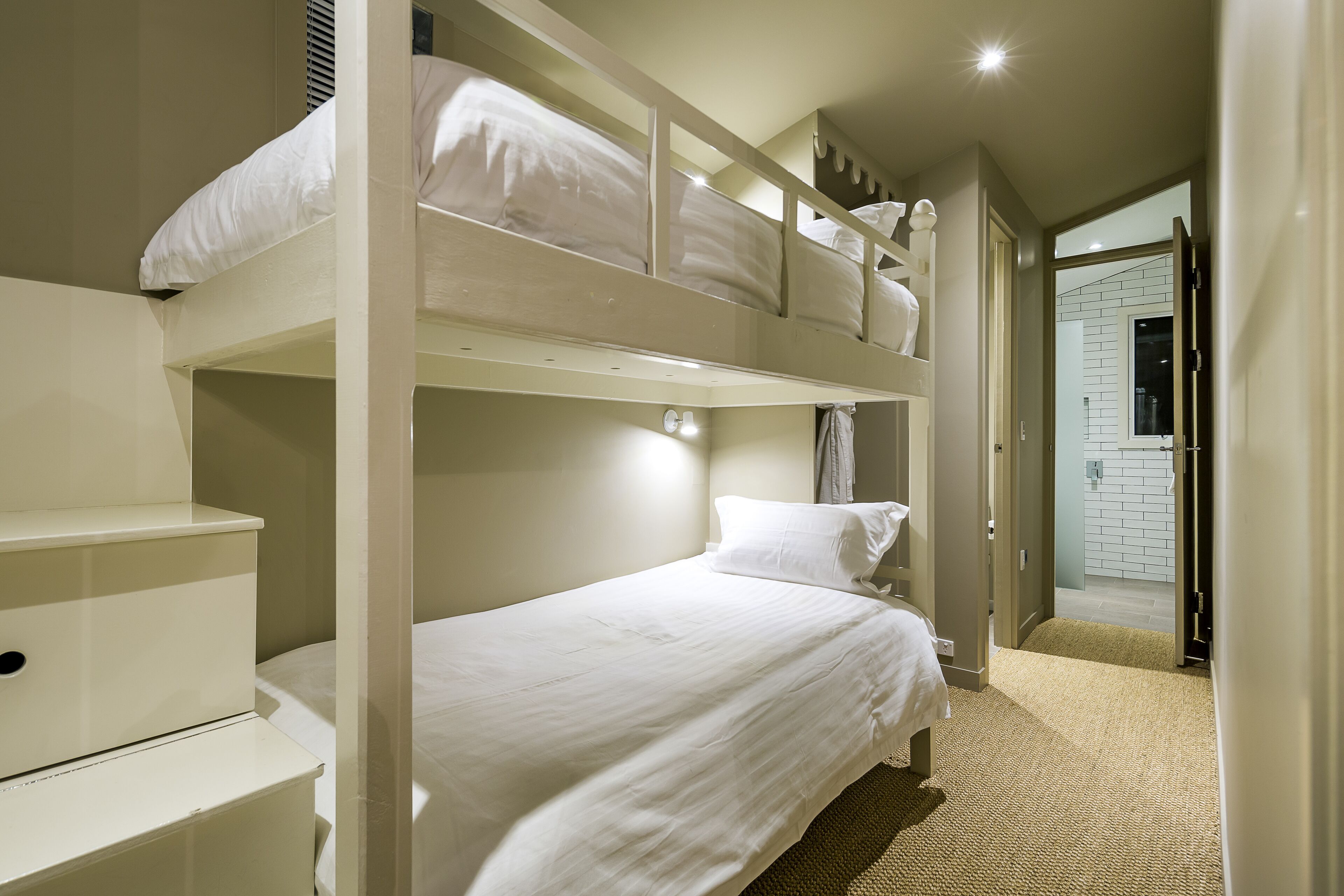 Habitación con literas, una paleta de colores blanca, una mesita de noche pequeña y una puerta que da a un baño.