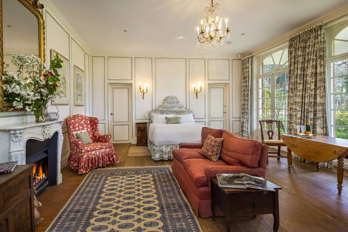 A spacious room with a large bed, a red armchair, a sofa, a wooden table, and a fireplace.