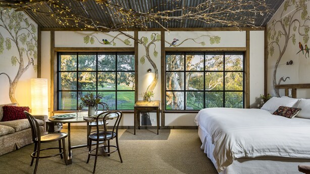 Ein Schlafzimmer mit einem großen Bett, einem Esstisch mit Stühlen und Blick auf Bäume durch große Fenster.