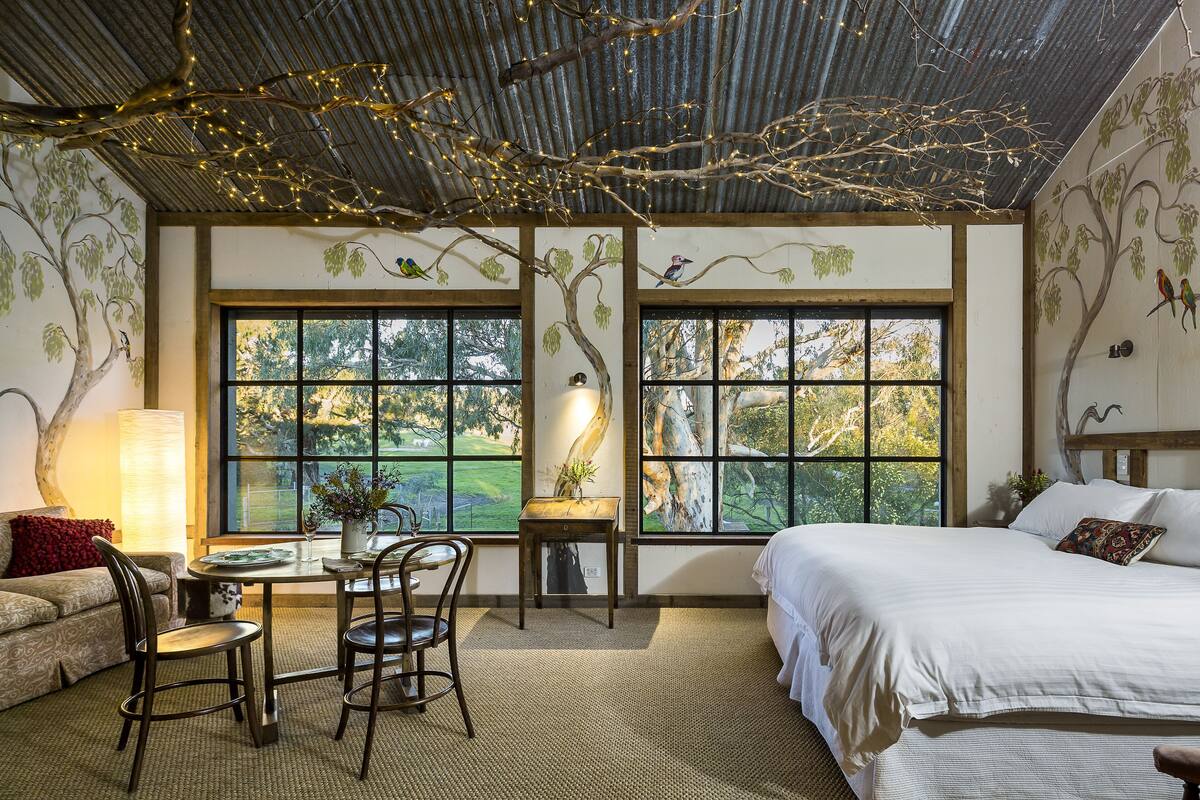 A bedroom with a large bed, a dining table with chairs, and a view of trees through large windows.