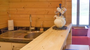 Une cuisine de style rustique, avec des éléments en bois, comprenant un évier, une théière et des tasses posées sur un sous-verre, ainsi qu’une fenêtre équipée de stores.