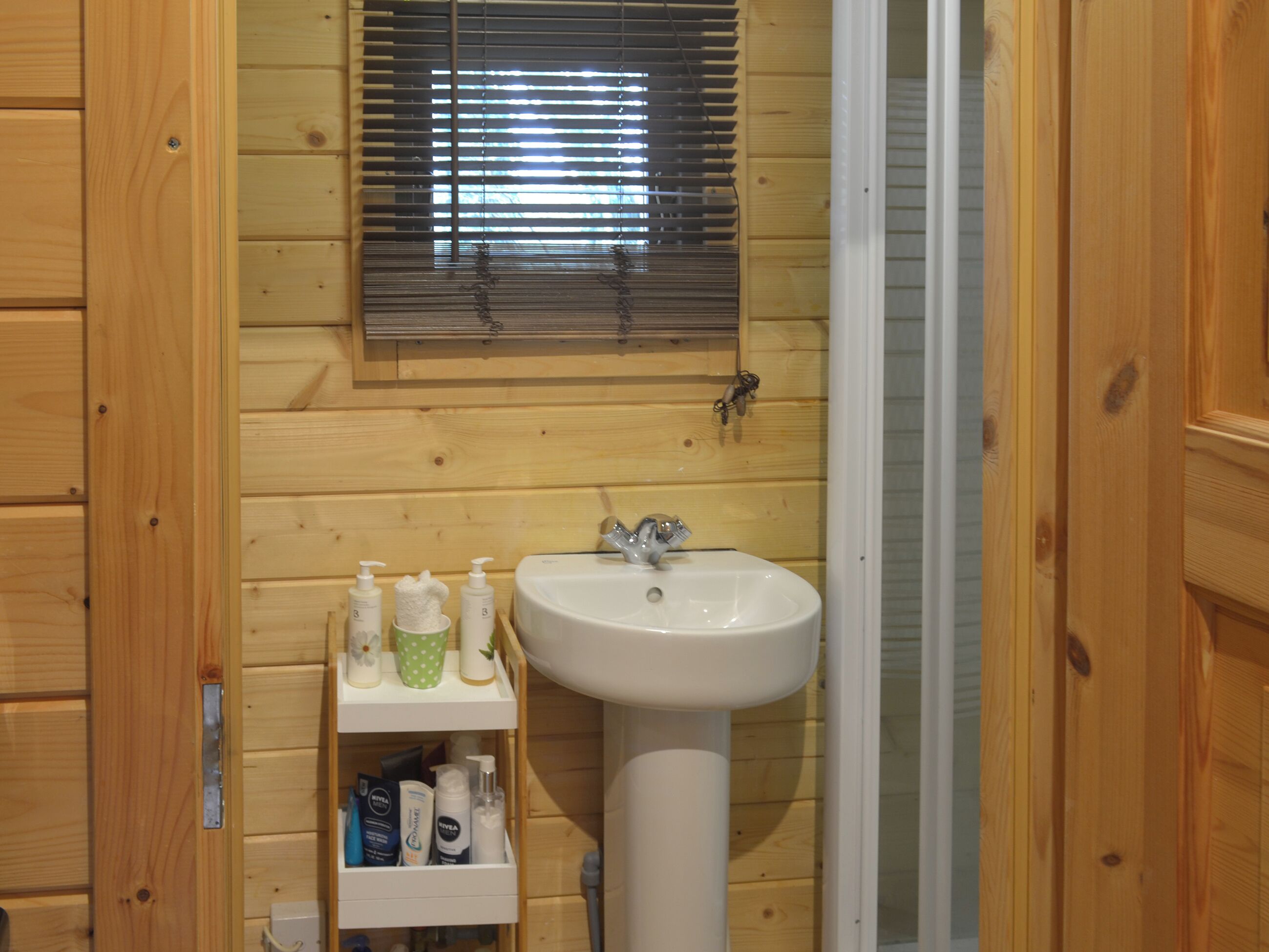 Une salle de bain en bois avec un lavabo, des stores aux fenêtres et des étagères contenant des articles de toilette.