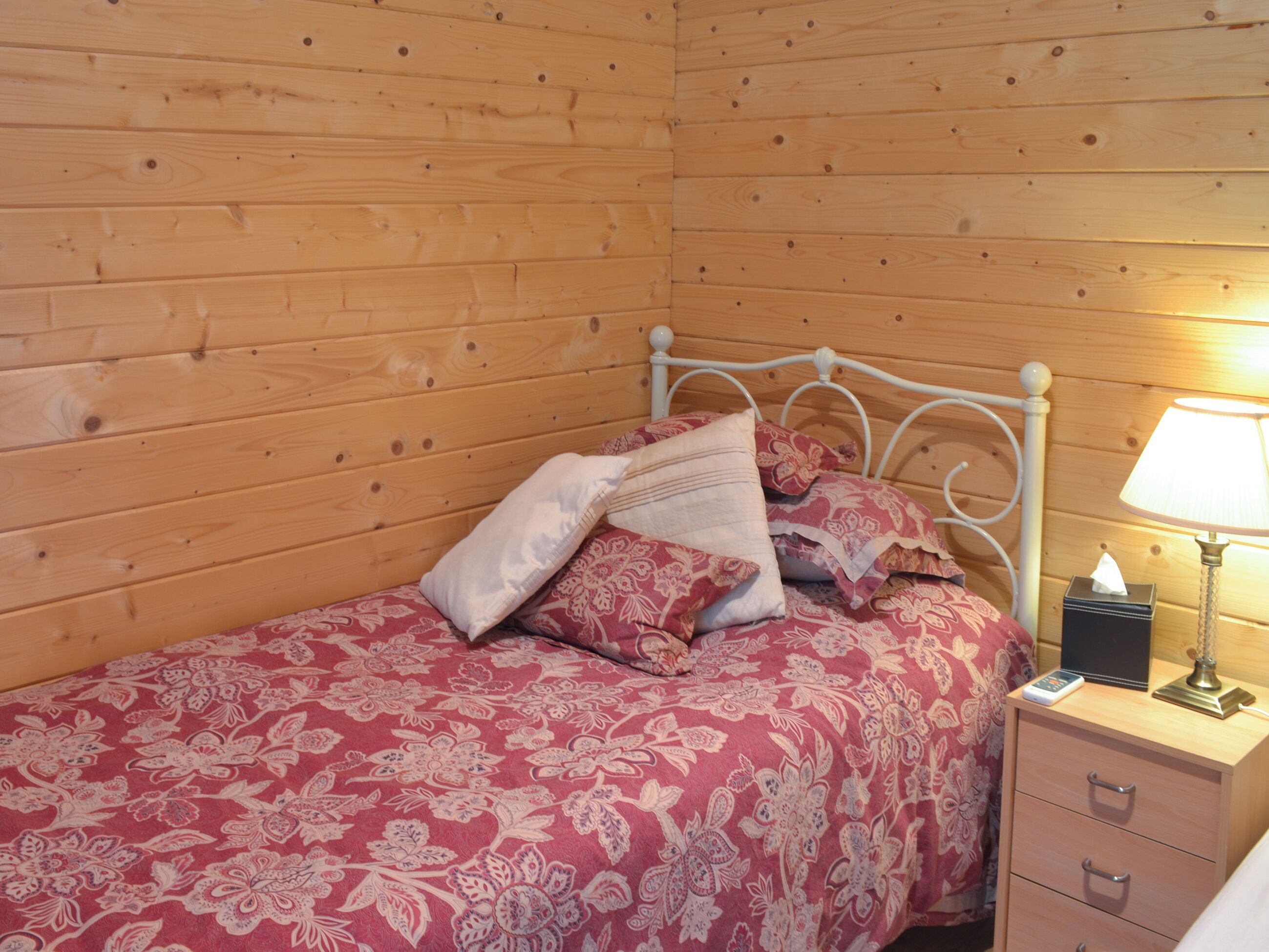 Ein Holzhütten-Zimmer mit einem Bett, floralem Bettzeug, einer Nachttischlampe und einem Nachttisch.