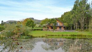 A house with a pond in front, surrounded by trees and a hill in the background.