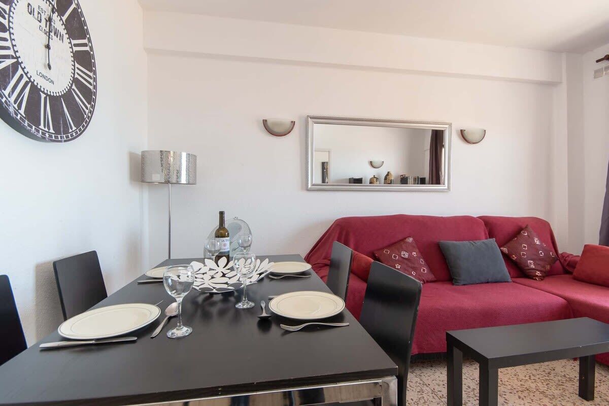 A dining area with a table set for two, a red sofa, and a wall-mounted clock.