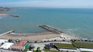 A coastal view with a beach, rocky breakwater, and a road leading to a parking area. - Radar Cabo Corrientes (Mar del Plata)