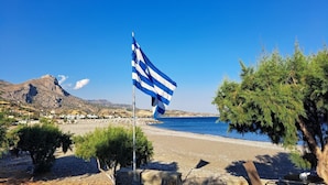 Una playa con un asta de banderas ondeando un pendón, un cielo azul despejado y montañas al fondo.