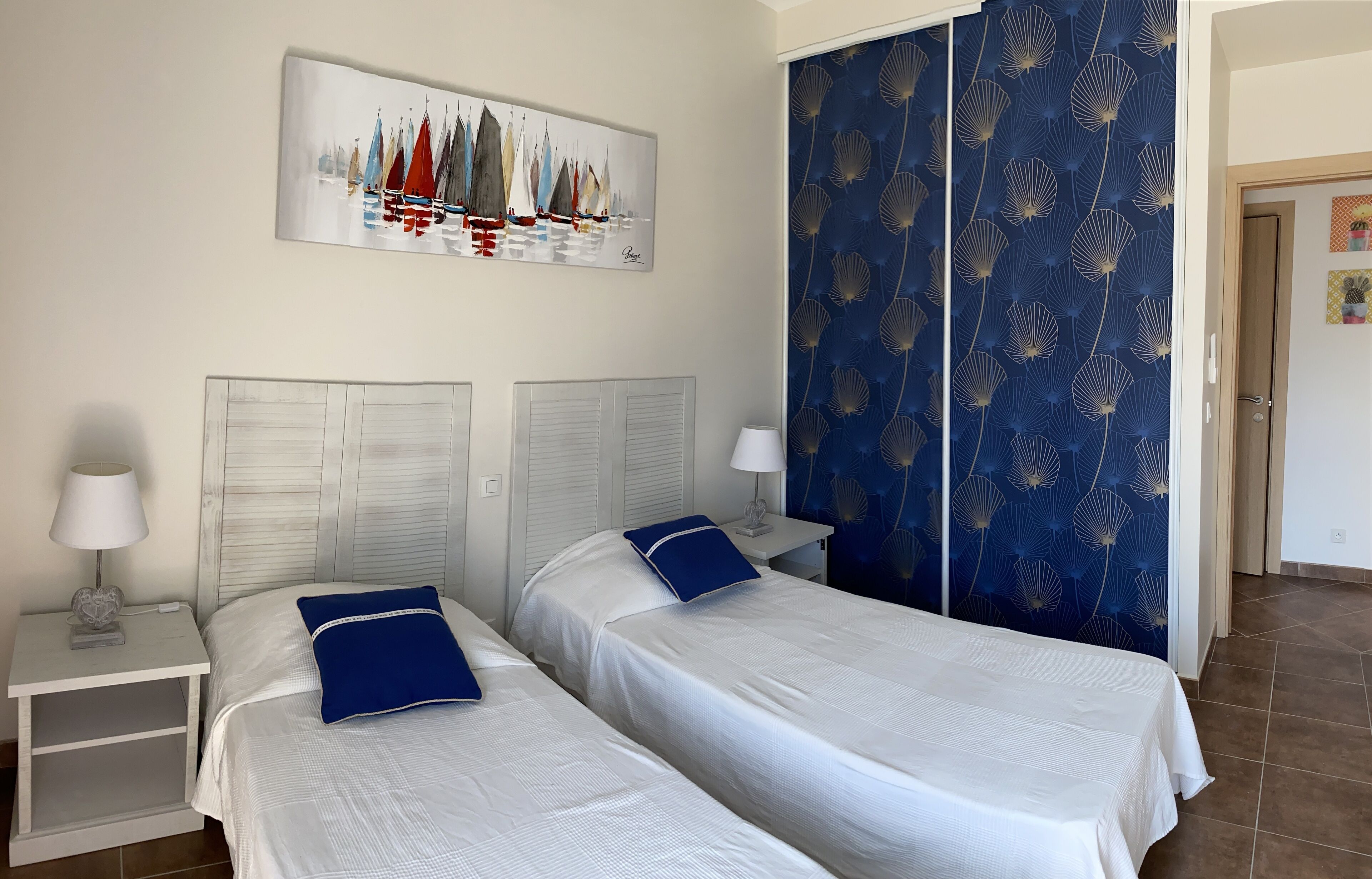 A hotel room with two beds, a painting of sailboats, a blue patterned wall, and a door with a small picture.
