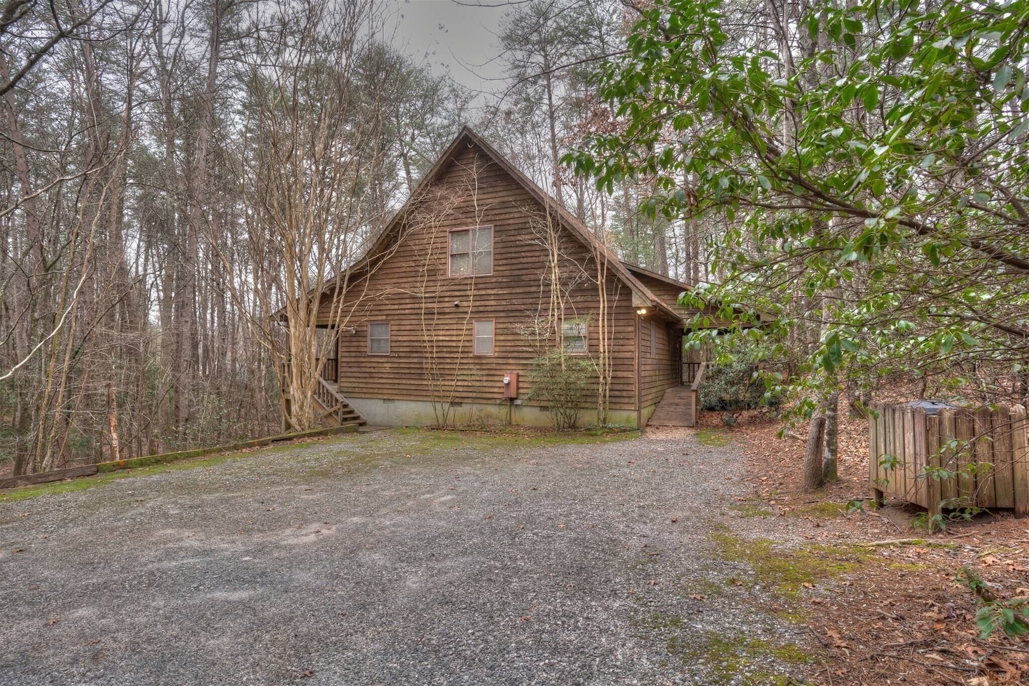 A rustic wooden cabin with a gravel driveway, surrounded by dense forest.