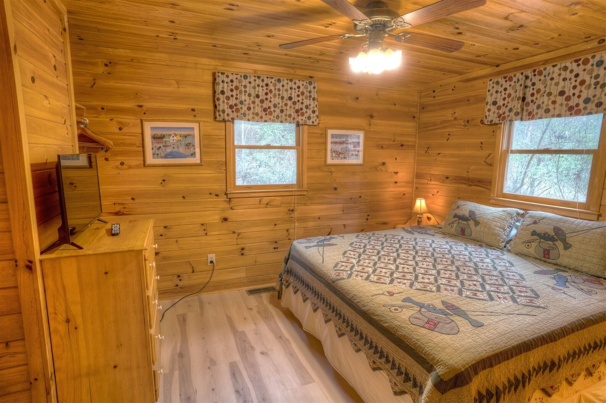 A wooden cabin bedroom with a bed, two windows, a ceiling fan, and a dresser.