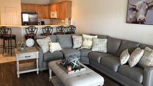 Un salon moderne avec un canapé d’angle gris, une table basse et un coin repas doté de meubles de rangement en bois et d’un réfrigérateur.