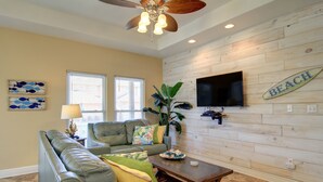 A living room with a sectional sofa, a coffee table, a ceiling fan, a TV mounted on a wooden wall, and a 'BEACH' sign.