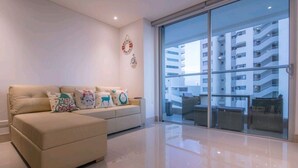 Una sala de estar moderna con un sofá color beige, almohadones decorativos y un amplio ventanal que da a un balcón con vistas a un edificio de gran altura.
