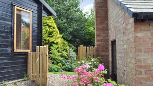 A black wooden building with a window and a brick wall. There is a wooden fence and a garden with pink flowers. - 2 bedroom accommodation in Tibthorpe, near Driffield (Tibthorpe, near Driffield)