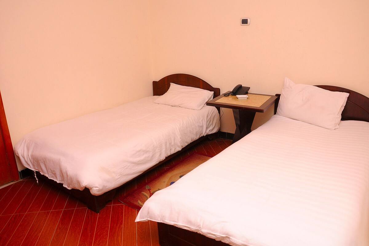 A room with two single beds, a wooden nightstand with a telephone, and a red tiled floor.