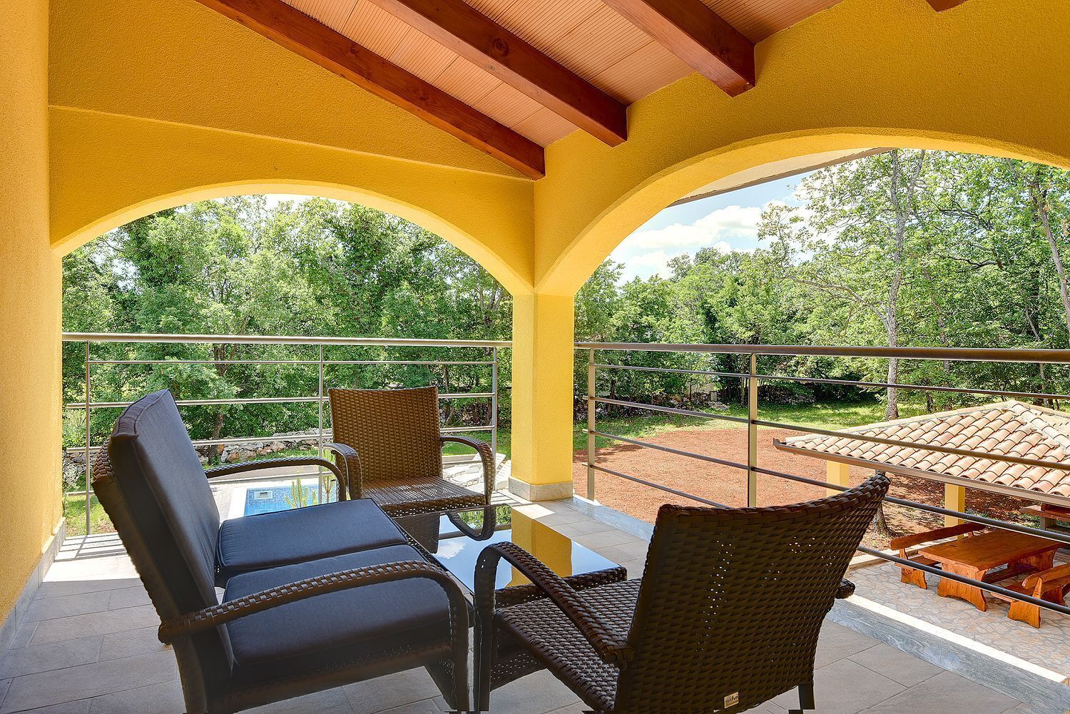 A covered outdoor seating area with wicker chairs and a table, overlooking a pool and greenery.