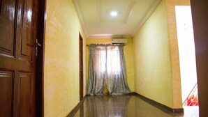 A hallway with a wooden door, yellow walls, a shiny floor, and a window with curtains. - White Rose Suites (Lagos)