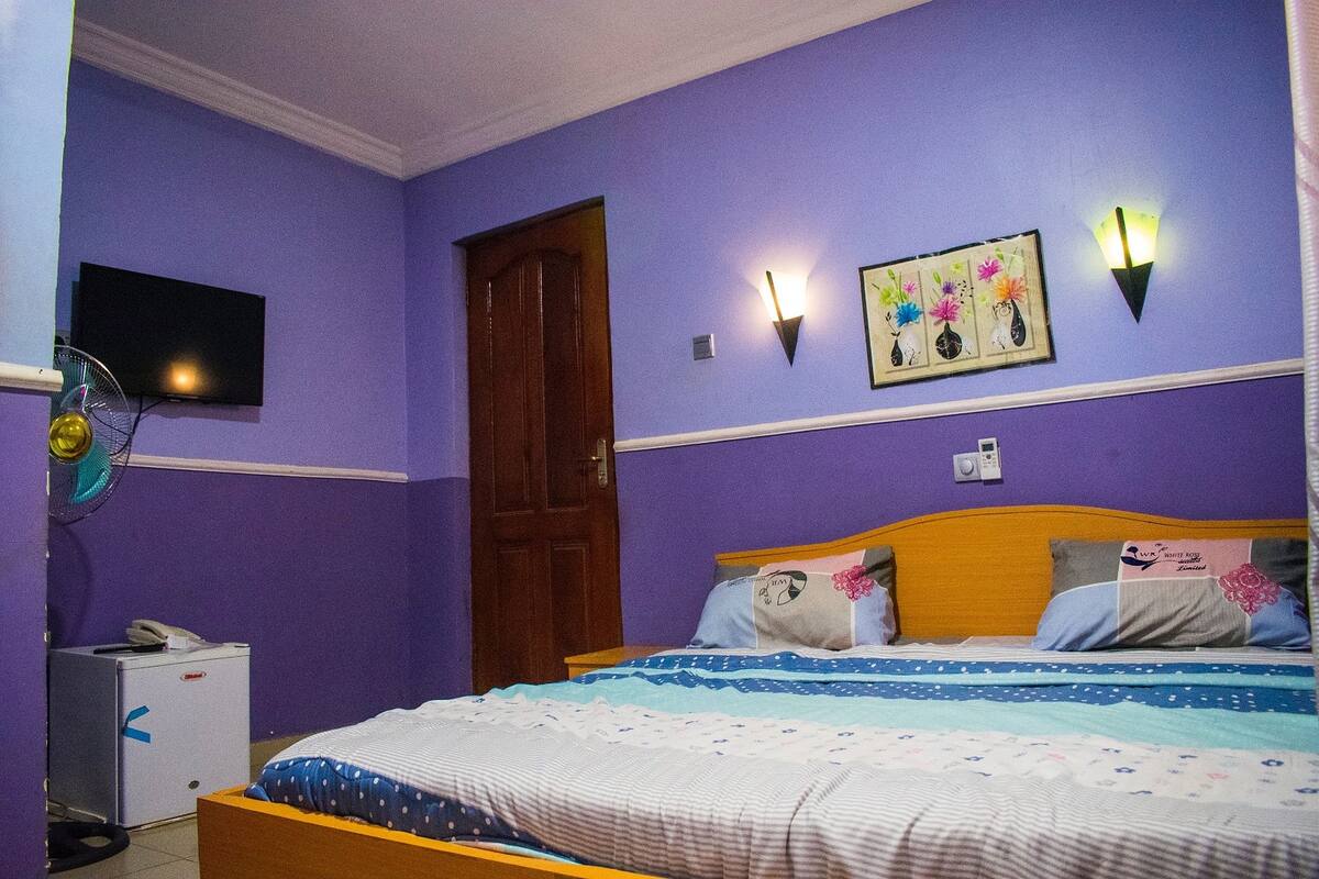 A bedroom with a bed, a television, a wall-mounted light, and a small refrigerator.