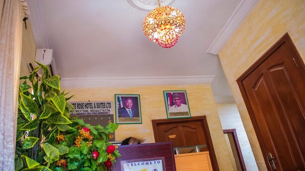 A reception area with a reception desk, a large decorative plant, and framed portraits on the wall. - White Rose Suites (Lagos)