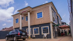 A two-story building with a patterned facade, a black SUV parked in front, and a small outdoor seating area. - White Rose Suites (Lagos)
