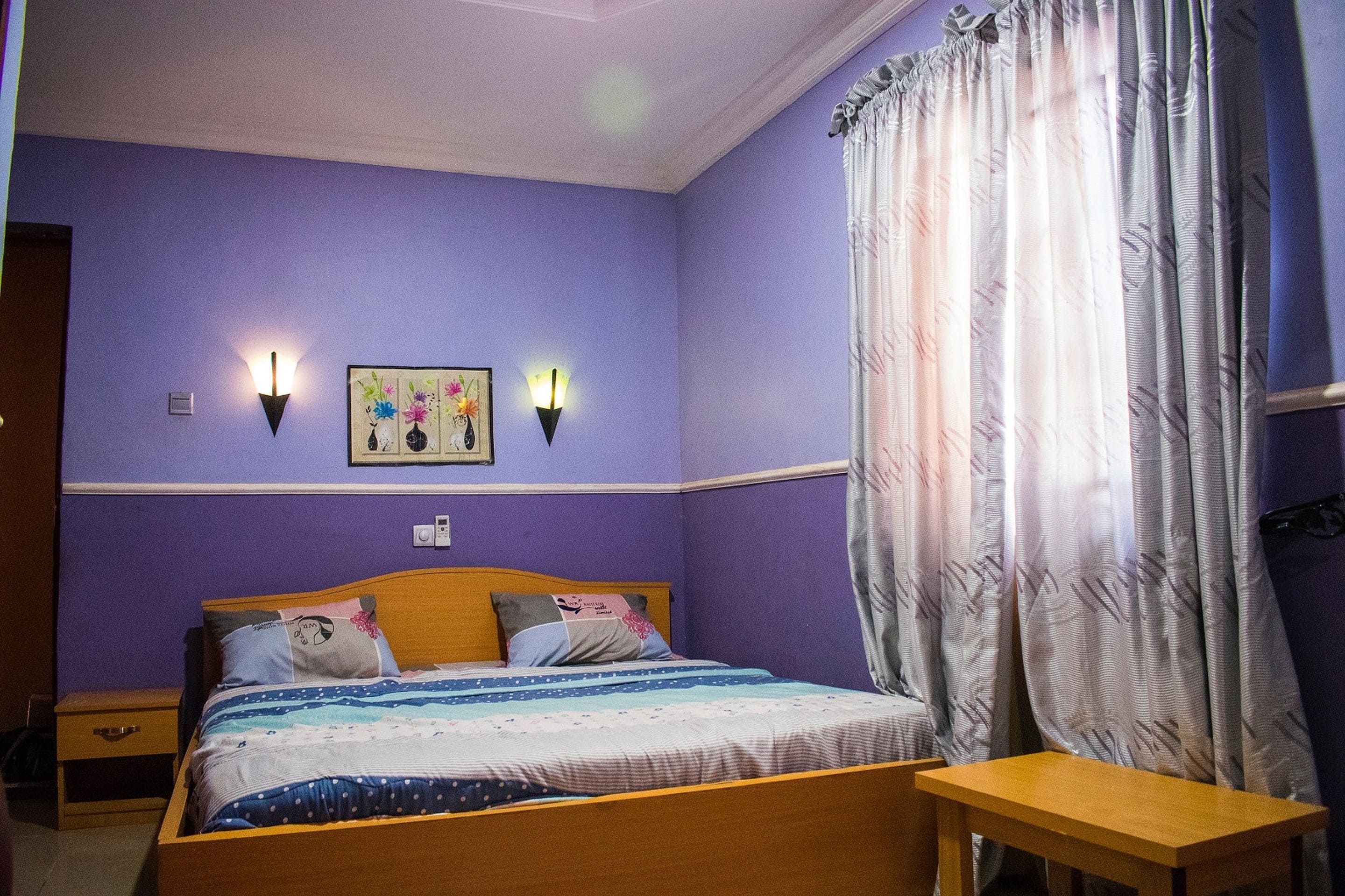 Un dormitorio con cama, mesitas de noche, lámpara de pared y ventana con cortinas.