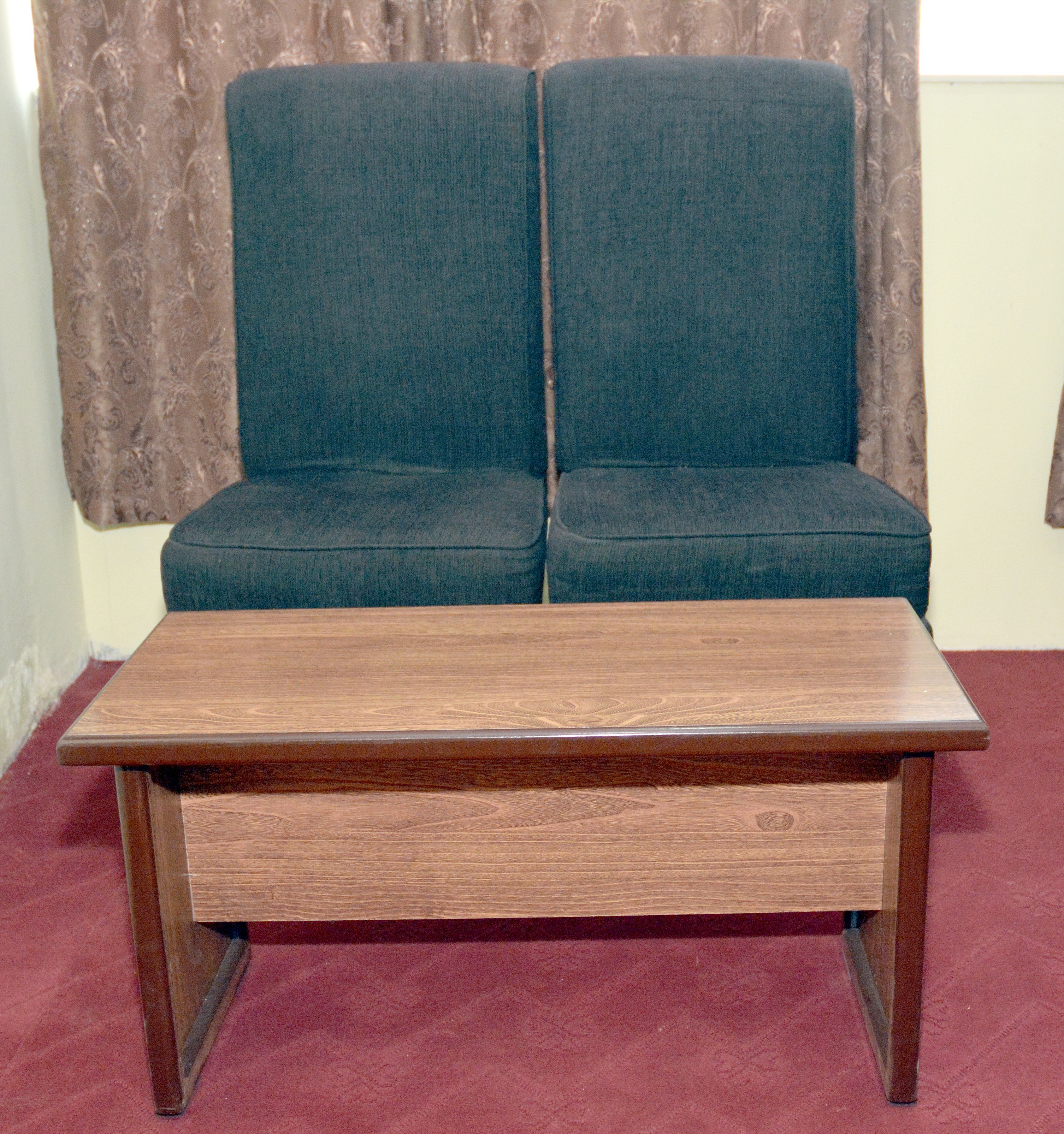 A wooden coffee table with a drawer in front of a blue sofa set against a patterned curtain.