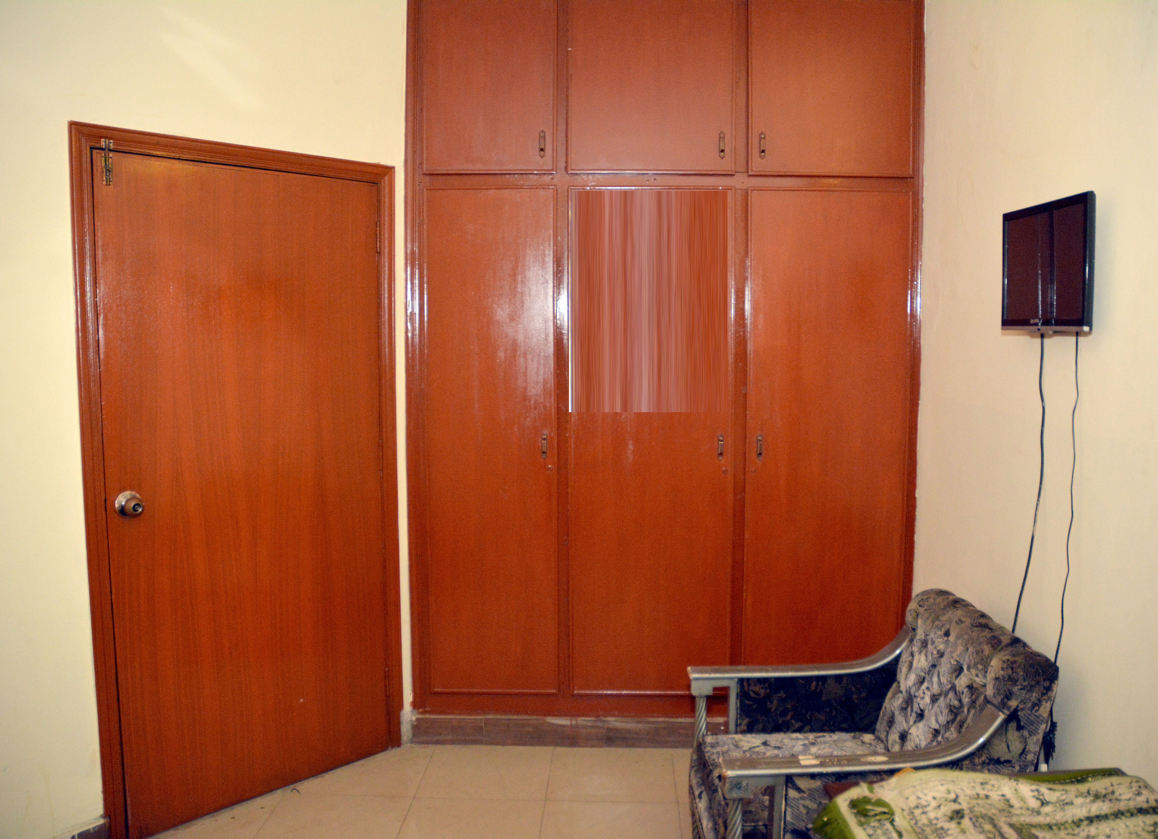 A room with a wooden wardrobe, a small table with a patterned tablecloth, and a wall-mounted TV.