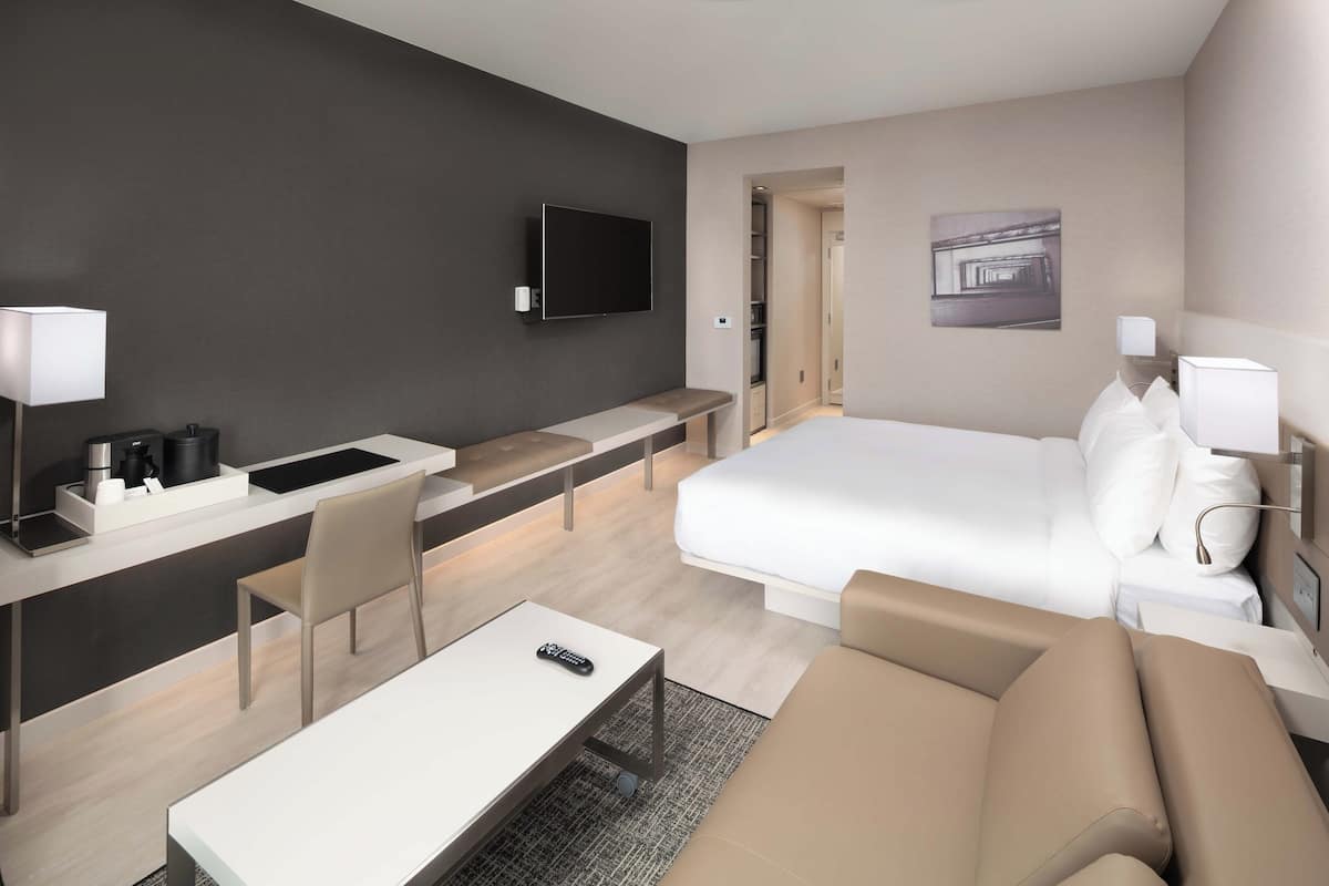 A modern hotel room with a bed, a desk with a chair, a TV mounted on the wall, and a bedside table with a lamp.