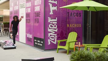 A vibrant outdoor seating area with green chairs and a table, a purple wall with 'Zombie Taco' branding, and a person walking by.