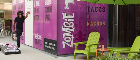 Et livligt udendørs siddeområde med grønne stole og et bord, en lilla væg med 'Zombie Taco' branding, og en person der går forbi.