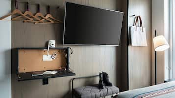 Une chambre d’hôtel moderne équipée d’un téléviseur à écran plat, d’un panneau d’affichage où l’on peut épingler des photos et des notes, d’un banc avec des bottes, d’un porte-manteau suspendu et d’une lampe de chevet.