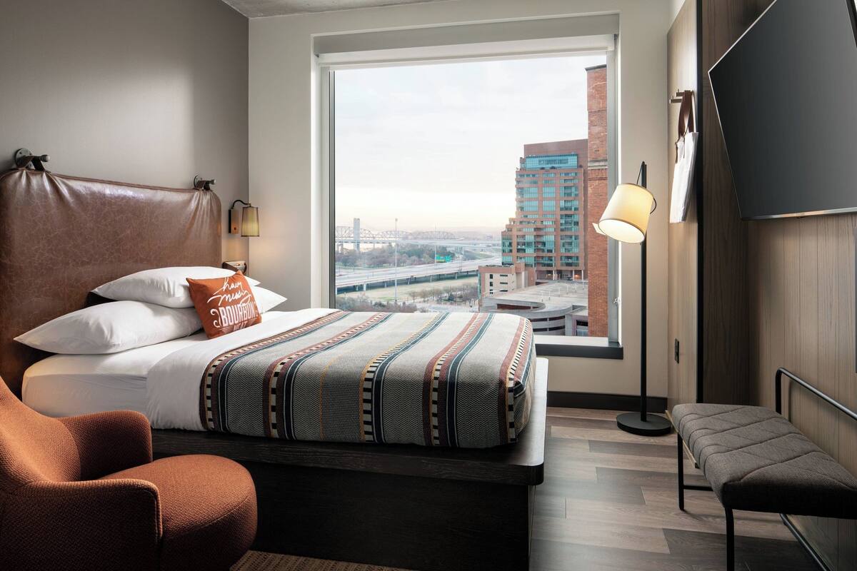 Une chambre d’hôtel moderne avec une grande fenêtre, un lit recouvert d’une courtepointe à motifs, une tête de lit en cuir marron, une lampe de chevet et une vue sur le paysage urbain.