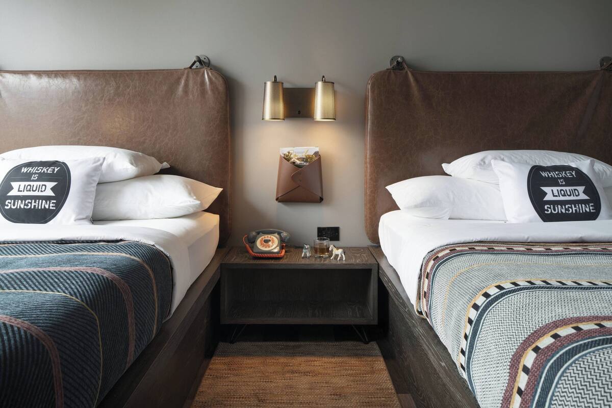 Une chambre d’hôtel avec deux lits, chacun orné d’un coussin sur lequel est brodé le texte : « Le whisky, c’est du soleil liquide. » On y trouve des lampes fixées au mur, une table de chevet avec un téléphone, un verre de whisky et une petite figurine.
