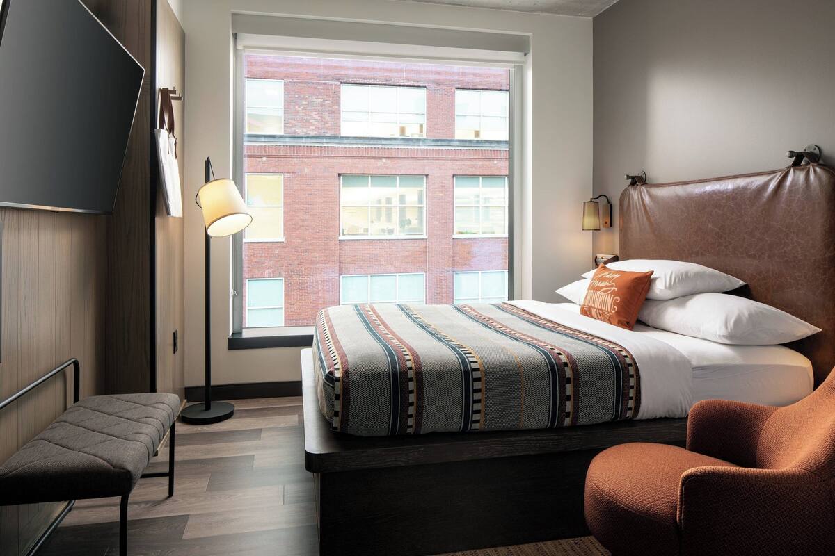 Une chambre d’hôtel moderne dotée d’une grande fenêtre, d’un lit avec une literie à motifs, d’une tête de lit en cuir, d’un banc et d’un lampadaire.