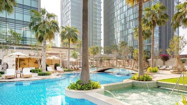 Un área de piscina moderna de hotel con una piscina cristalina, sillas de descanso, palmeras y un puente.