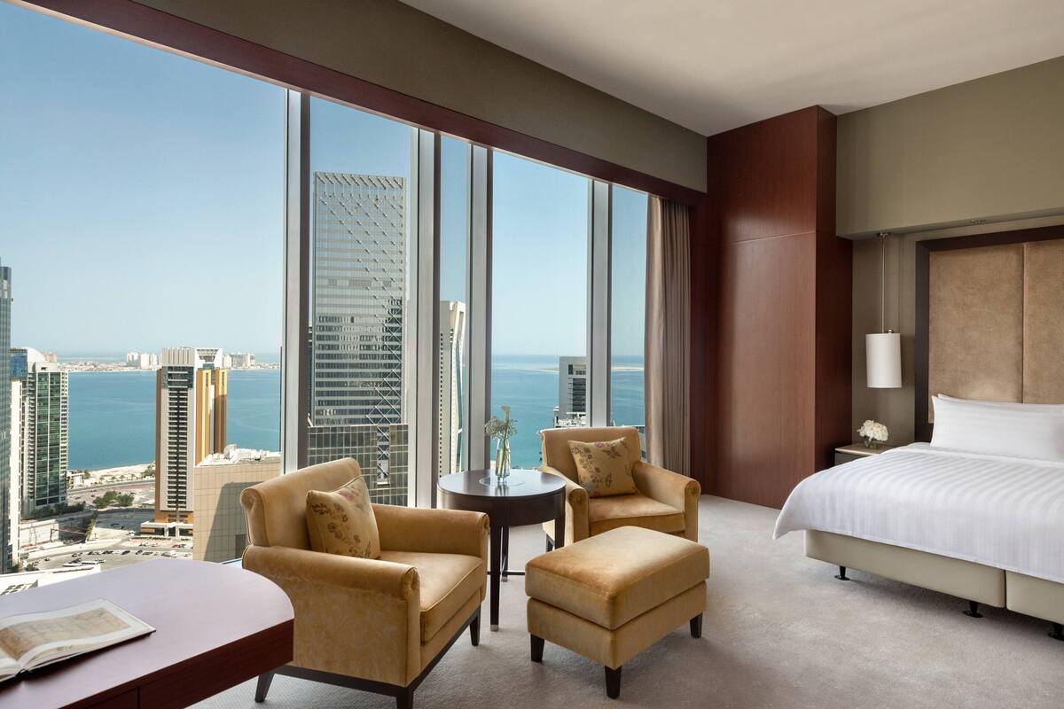 A hotel room with a large window, a bed, two armchairs, a small table, and a view of the city and water.
