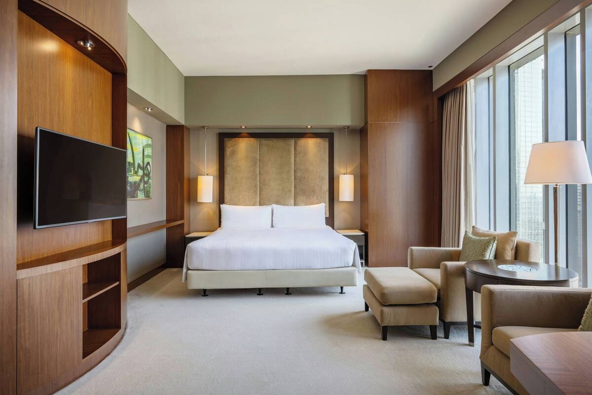 A hotel room with a large bed, a flat-screen TV, a seating area with a table, and a large window with curtains.