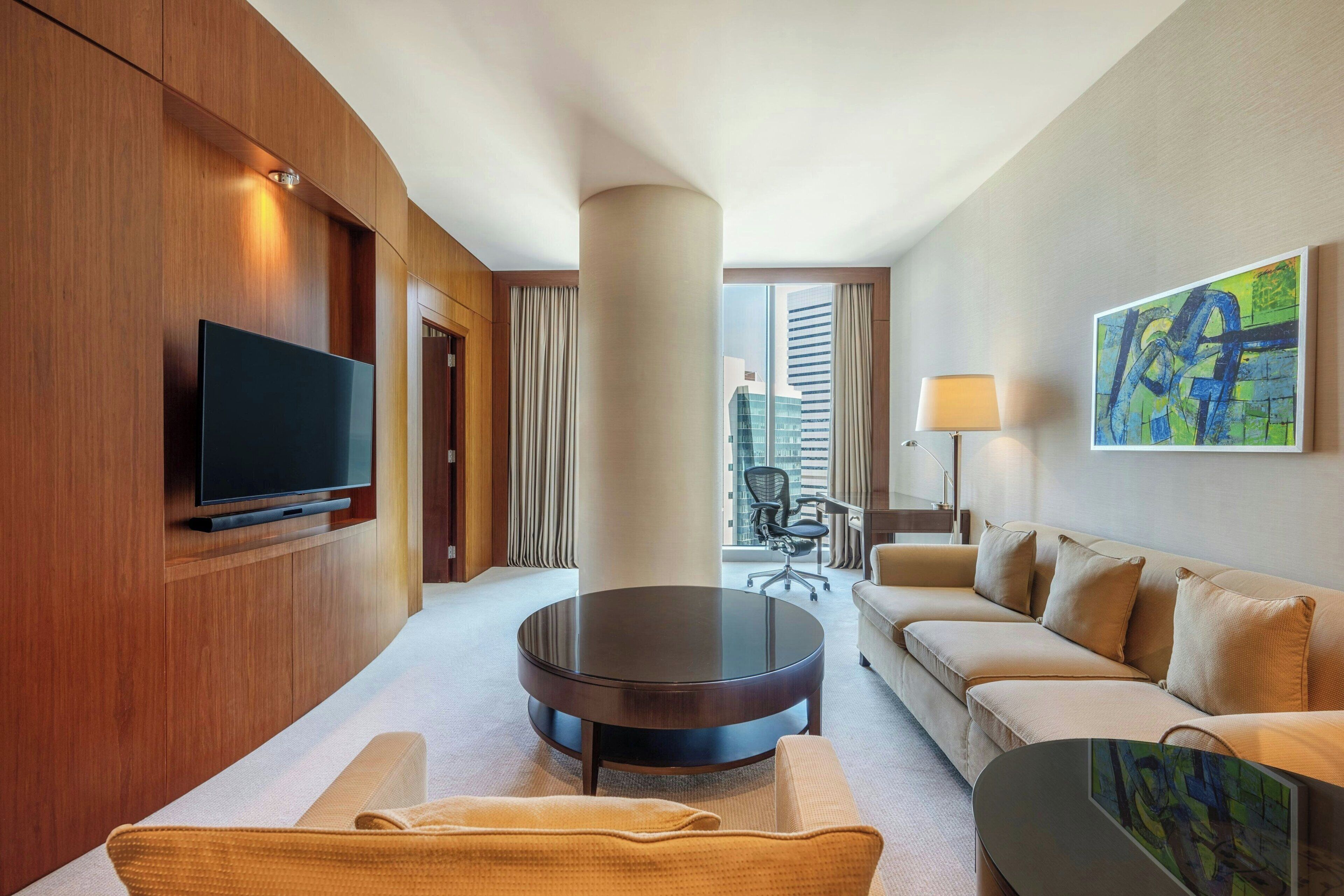 Una habitación de hotel moderna con una televisión de pantalla plana, una mesa de centro redonda, un sofá, una lámpara y un cuadro en la pared.