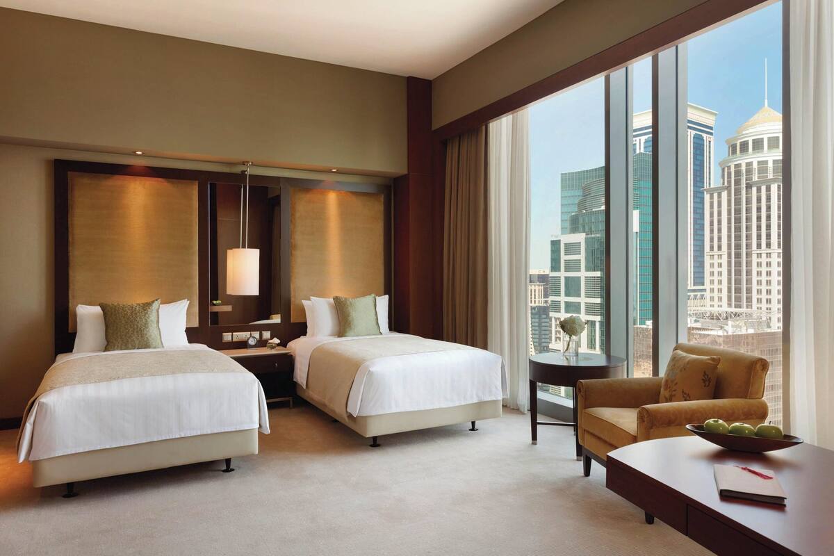 A hotel room with two beds, a desk, a chair, and a large window offering a city view.